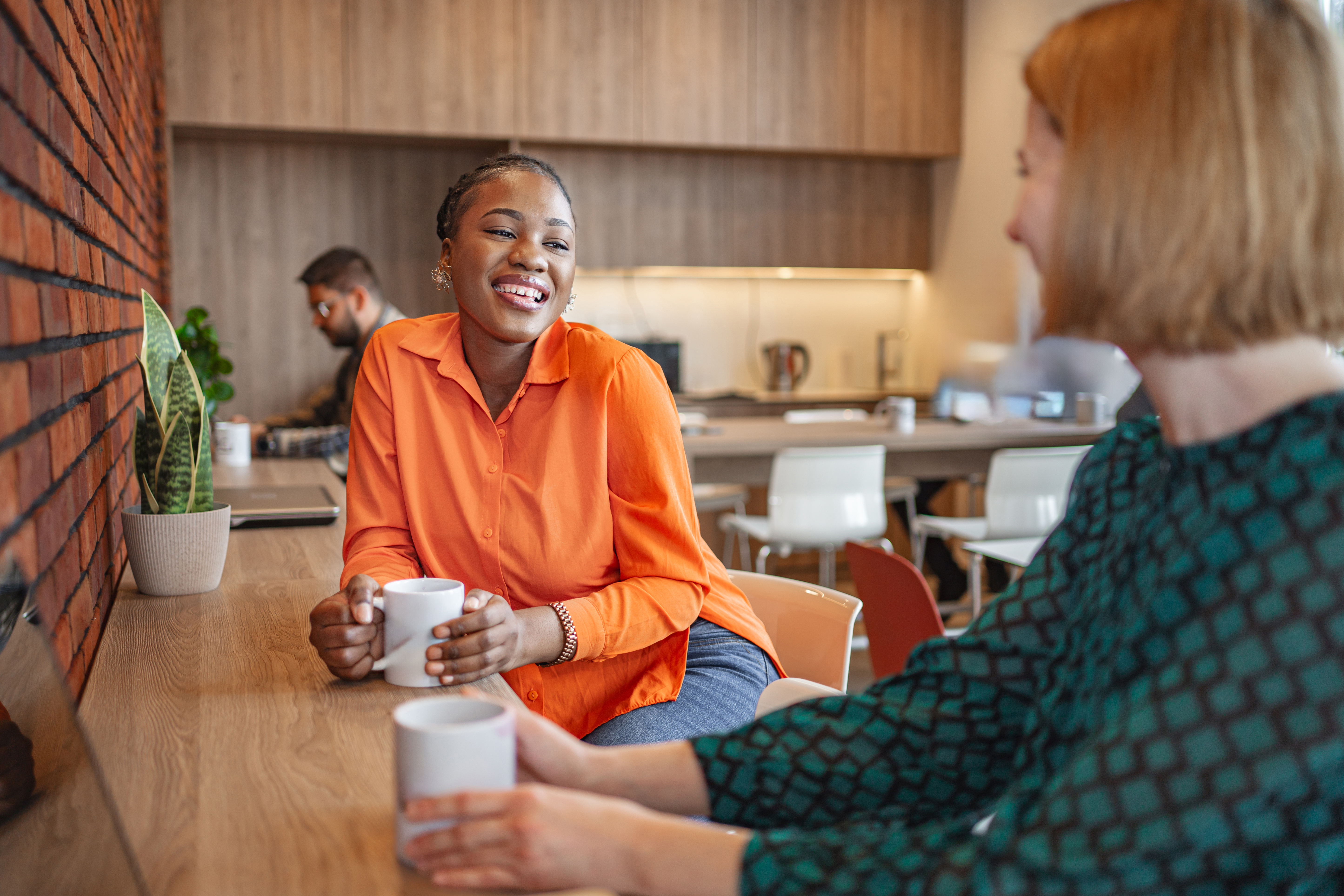 Two women seated at a high table in an office. They're holding coffee cups and appear to be chatting in a relaxed way.