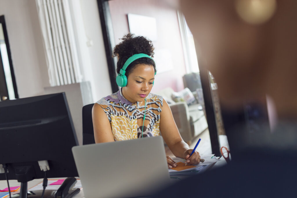 A woman wearing headphones and working in an office. She has a brightly coloured top and turqoise headphones.